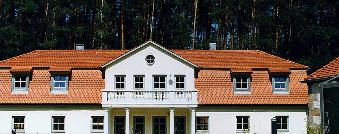 Weißes Mühlenanwesen mit rotem Ziegeldach vor Waldrand